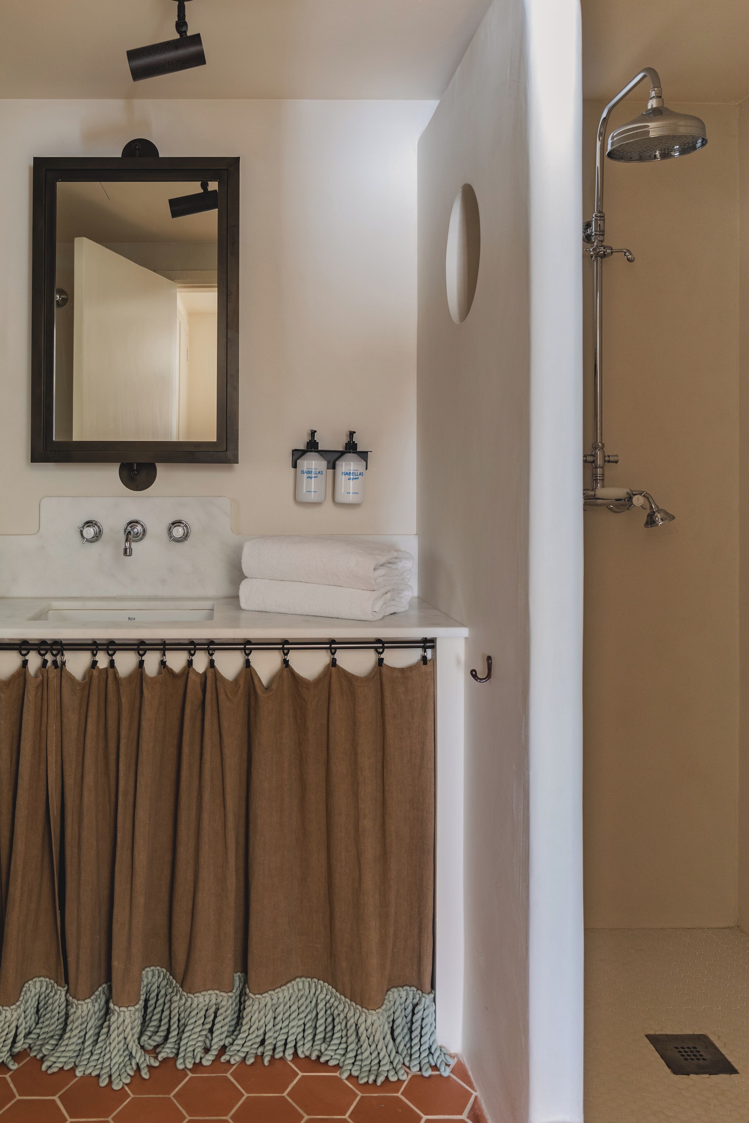 Baño con lavamanos blanco, espejo y ducha. Hay dos botellas de champú y acondicionador en la pared.