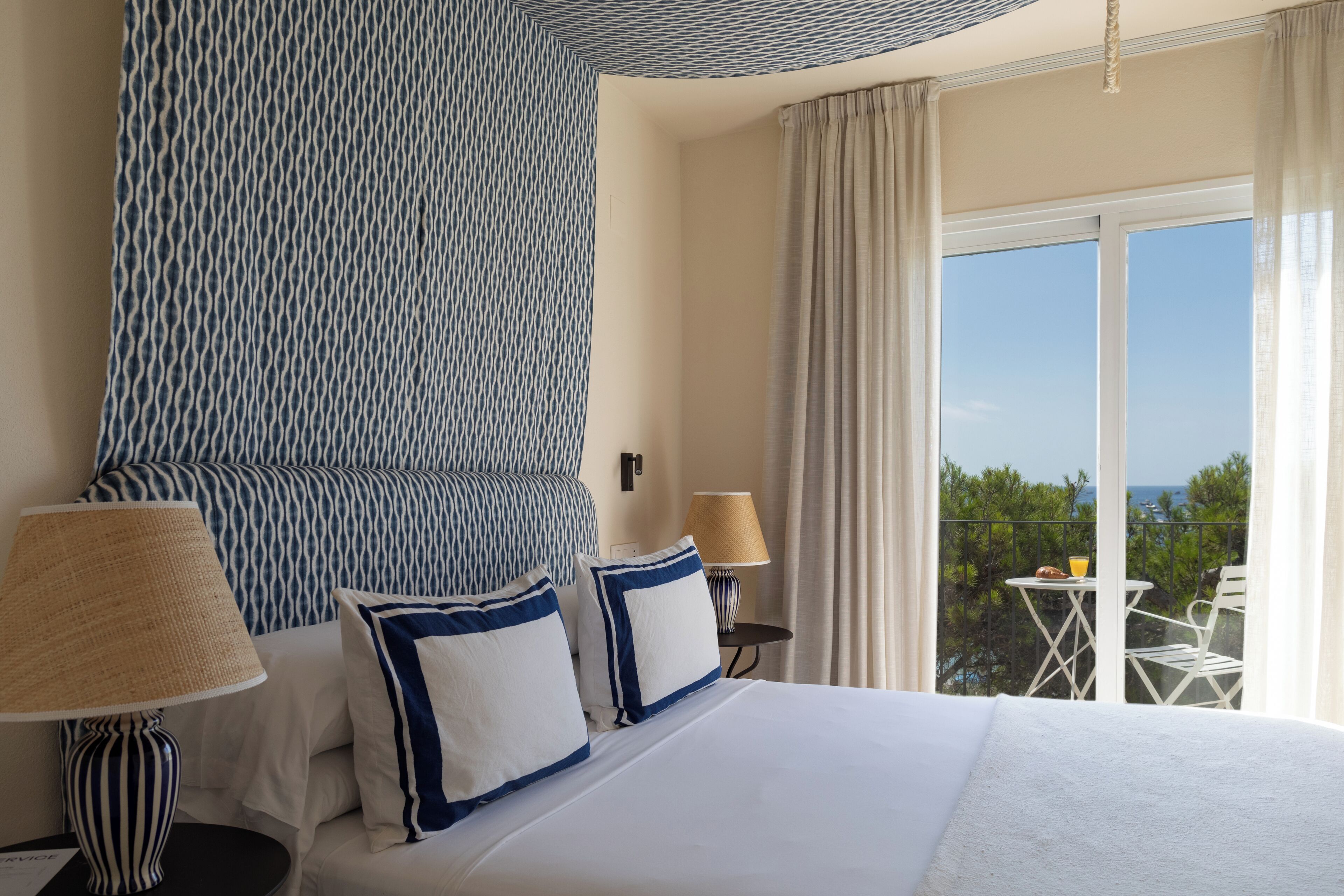 A hotel room with a bed, two bedside lamps, a small table with a chair, and a balcony with a table and chairs.