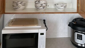 A kitchen with a microwave, a slow cooker, and a wooden cabinet with shelves holding plates and bowls.