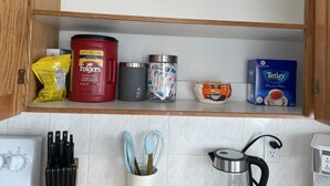A kitchen shelf with various items including a red Folgers coffee can, a grey container labeled 'TEA', a blue Tetley tea box, and a yellow packet.