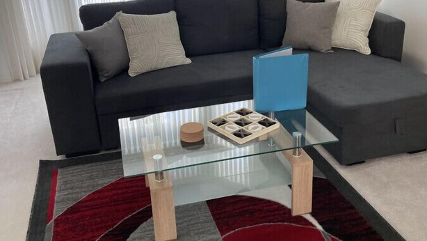 A modern living room with a dark grey sofa, two cushions, a glass coffee table, and a red and grey patterned rug.