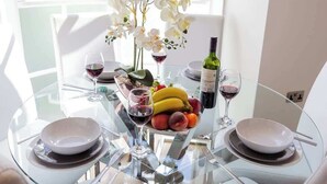 A round glass table with white plates, two wine glasses, a bottle of wine, a fruit bowl, and white orchids.