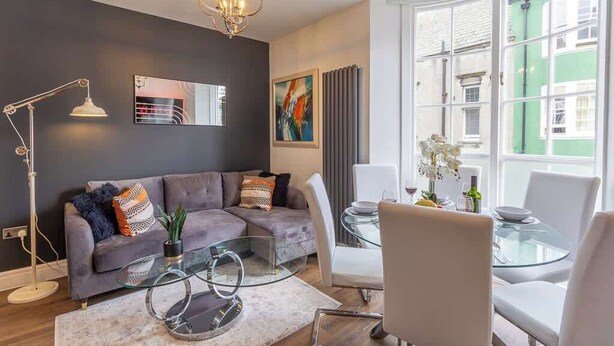 A modern living room with a grey sofa, a glass coffee table, and a dining area with a glass table and white chairs.