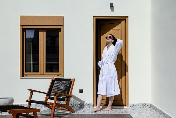 A person in a white bathrobe standing at a wooden door, with a wooden chair and a stone bench in the foreground. - Athens Feeling (Athens)