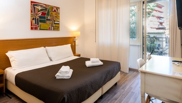 A hotel room with a bed, a desk, a chair, and a view of a traditional building outside. - Aurelia Home Guest House (Rome)