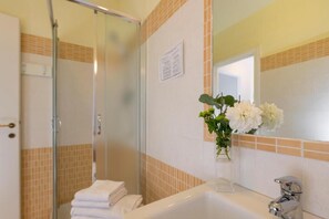 A bathroom with a glass shower enclosure, a white sink with a mirror, and a vase with white flowers. - Aurelia Home Guest House (Rome)