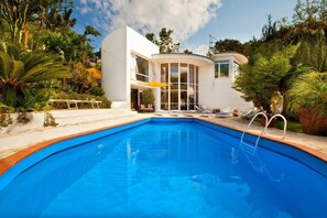 Une maison moderne avec une piscine, entourée d’une végétation luxuriante et sous un ciel dégagé.