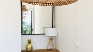 Un coin repas comprenant une table en bois, des chaises, un luminaire suspendu au plafond en fibres tressées, un miroir fixé au mur, une petite étagère avec un ananas et une lampe sur pied.