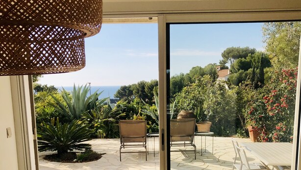 Un coin repas avec une table et des chaises, une lampe en osier et une vue sur un jardin et la mer, visible à travers une porte vitrée.