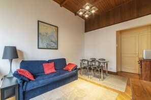 Un salon avec un canapé bleu, des coussins rouges, une table avec des chaises, une lampe et un tableau au mur.