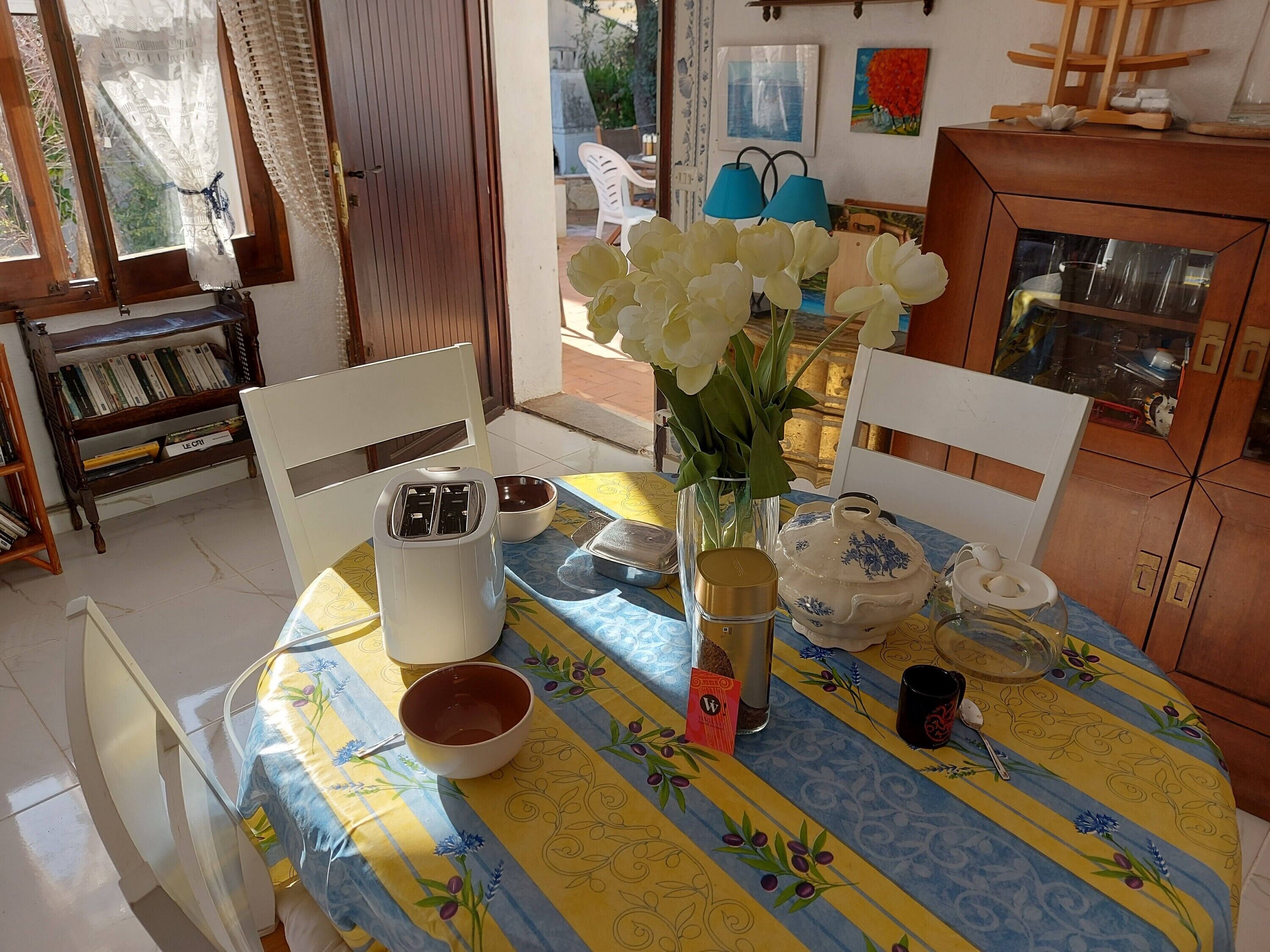 A dining area with a table set for two, featuring a yellow and blue floral tablecloth, a white toaster, a glass vase with white flowers, and a white teapot.