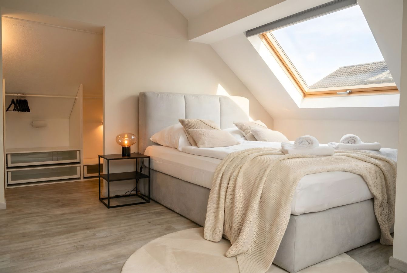 Ein Schlafzimmer mit Dachfenster, einem Bett mit Kissen, einem Nachttisch mit Lampe und einem Kleiderschrank mit Kleidung.