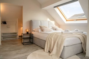 Ein Schlafzimmer mit Dachfenster, einem Bett mit Kissen, einem Nachttisch mit Lampe und einem Kleiderschrank mit Kleidung.