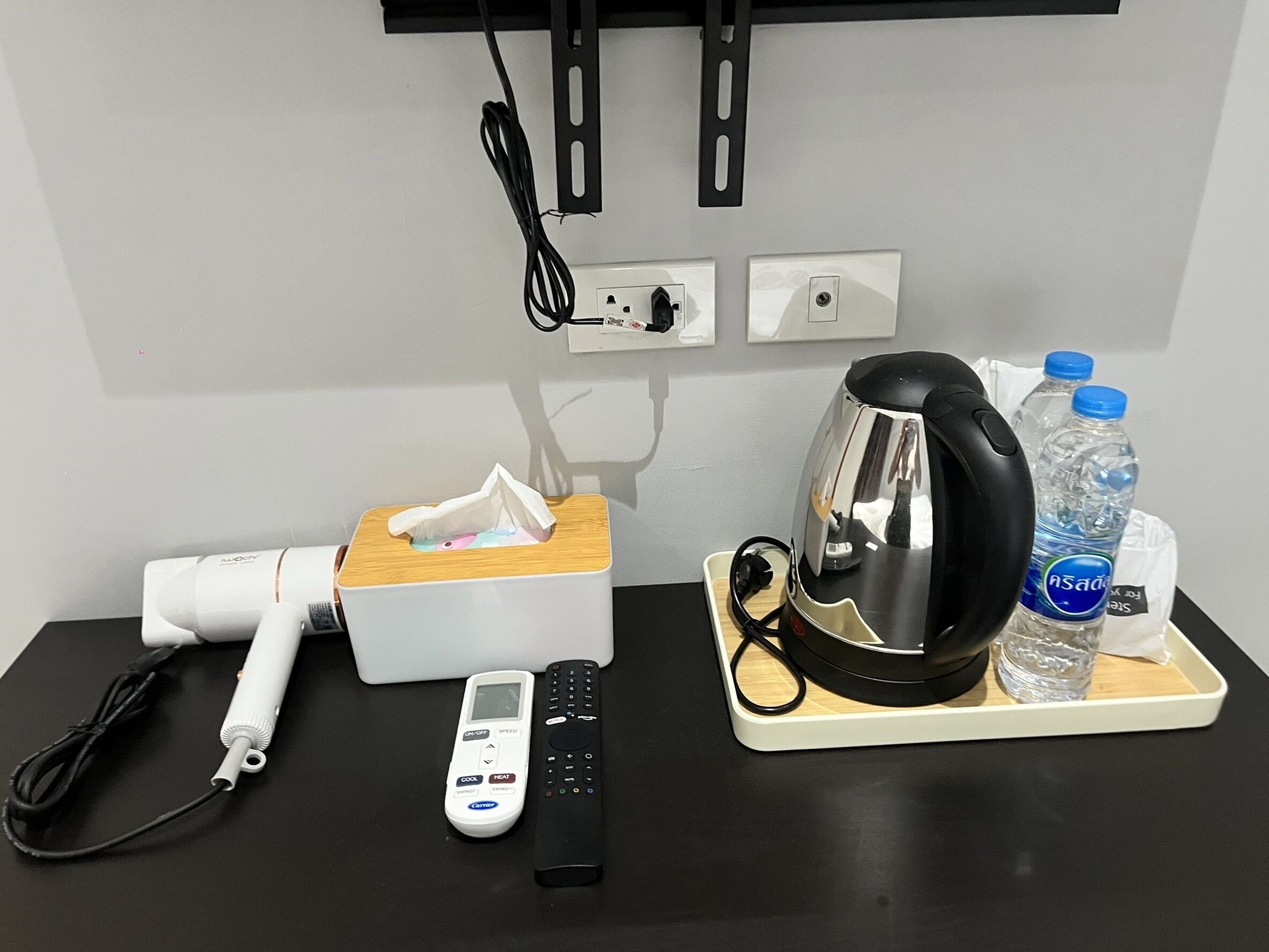 A hotel room with a desk, a tissue box, a hair dryer, a kettle, a remote control, and two water bottles.