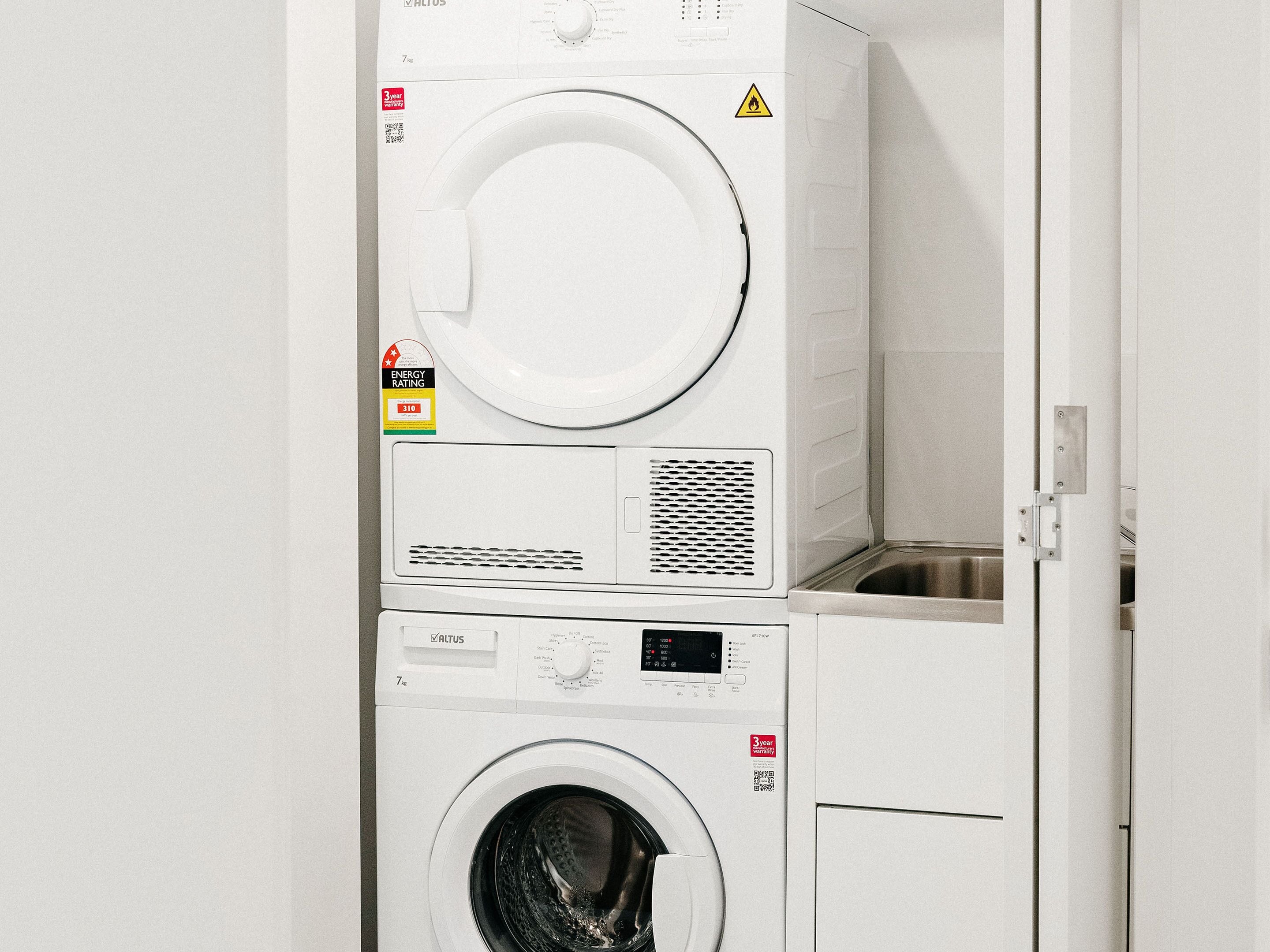 A stacked washer and dryer unit with a 3-year warranty label, energy rating sticker, and a caution warning sign.