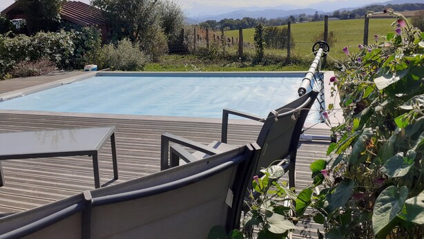 Una piscina con cubierta de madera y sillas, rodeada de vegetación y vistas a colinas distantes.