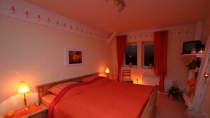 A bedroom with a large bed, two bedside lamps, a nightstand, a window with curtains, a radiator, and a television on a shelf. - Eckhorst Farm, Tuscany (Güby)