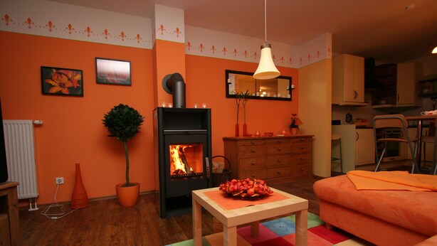 A living room with a fireplace, orange walls, a wooden table, and a colorful rug. - Eckhorst Farm, Tuscany (Güby)