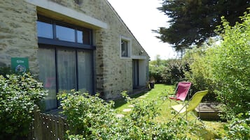 A stone building with a large window and a smaller window, surrounded by lush greenery and a wooden fence.
