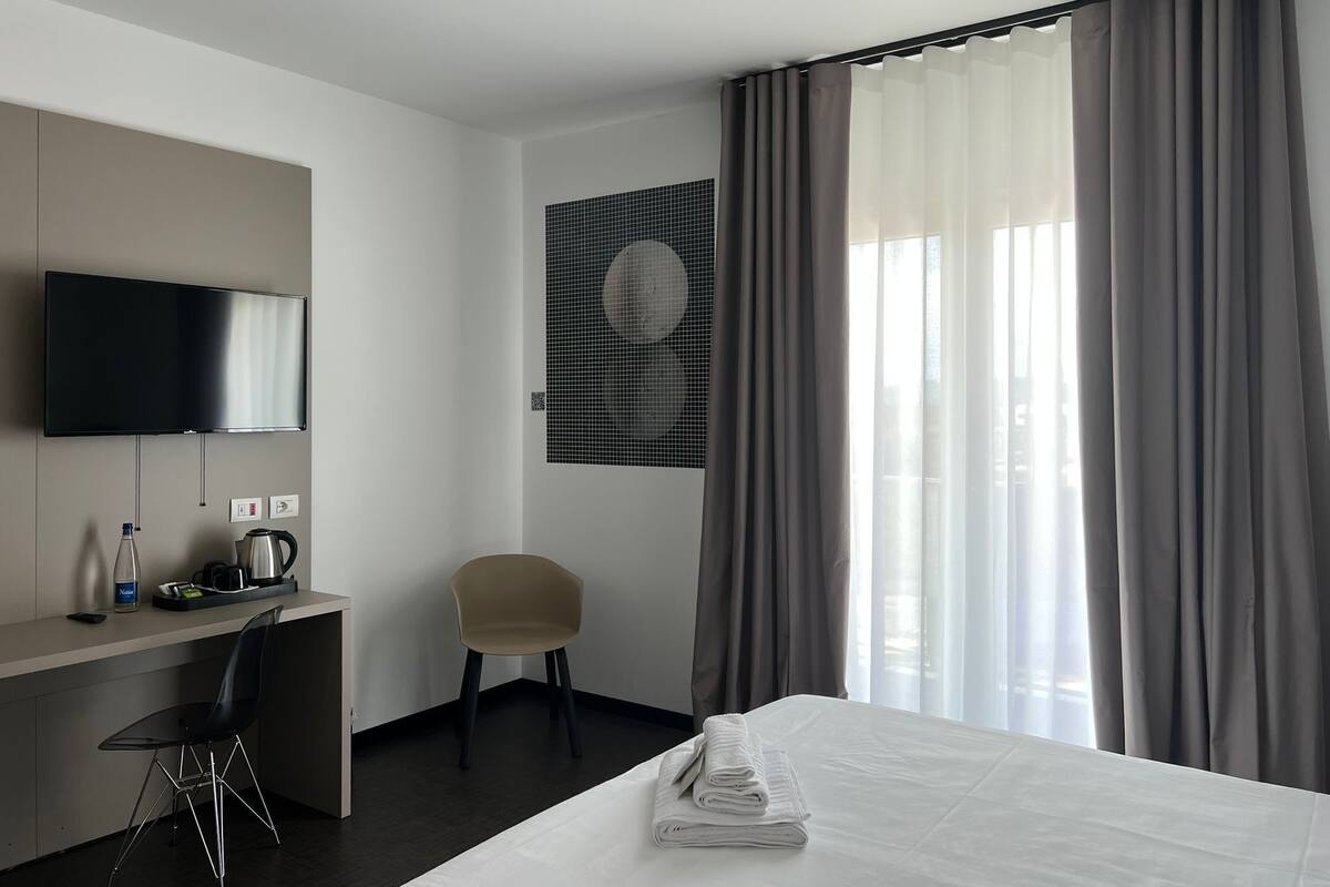 Ein Hotelzimmer mit einem Bett, einem Schreibtisch mit Stuhl, einem Fernseher und einem großen Fenster mit Vorhängen.