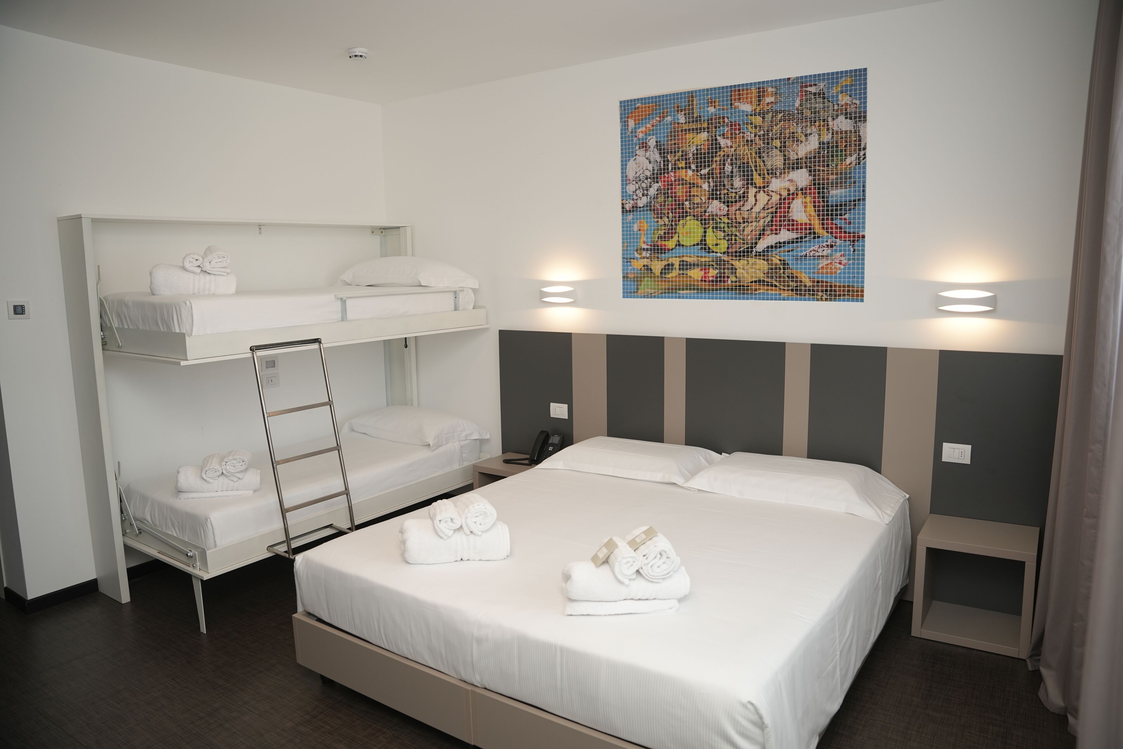 A hotel room with a bunk bed, a lower bunk with towels, a large bed with towels, a mosaic artwork on the wall, and bedside lamps.