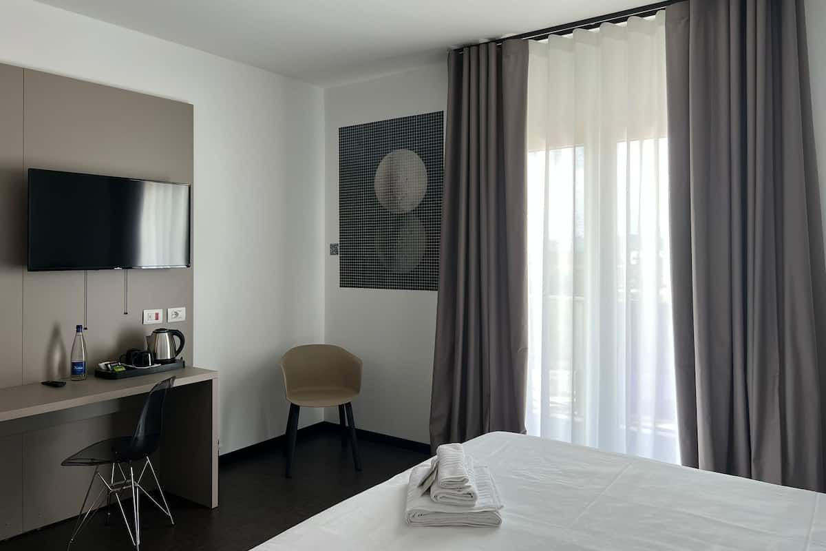 Ein Hotelzimmer mit einem Bett, einem Schreibtisch mit Stuhl, einem Fernseher und einem großen Fenster mit Vorhängen.