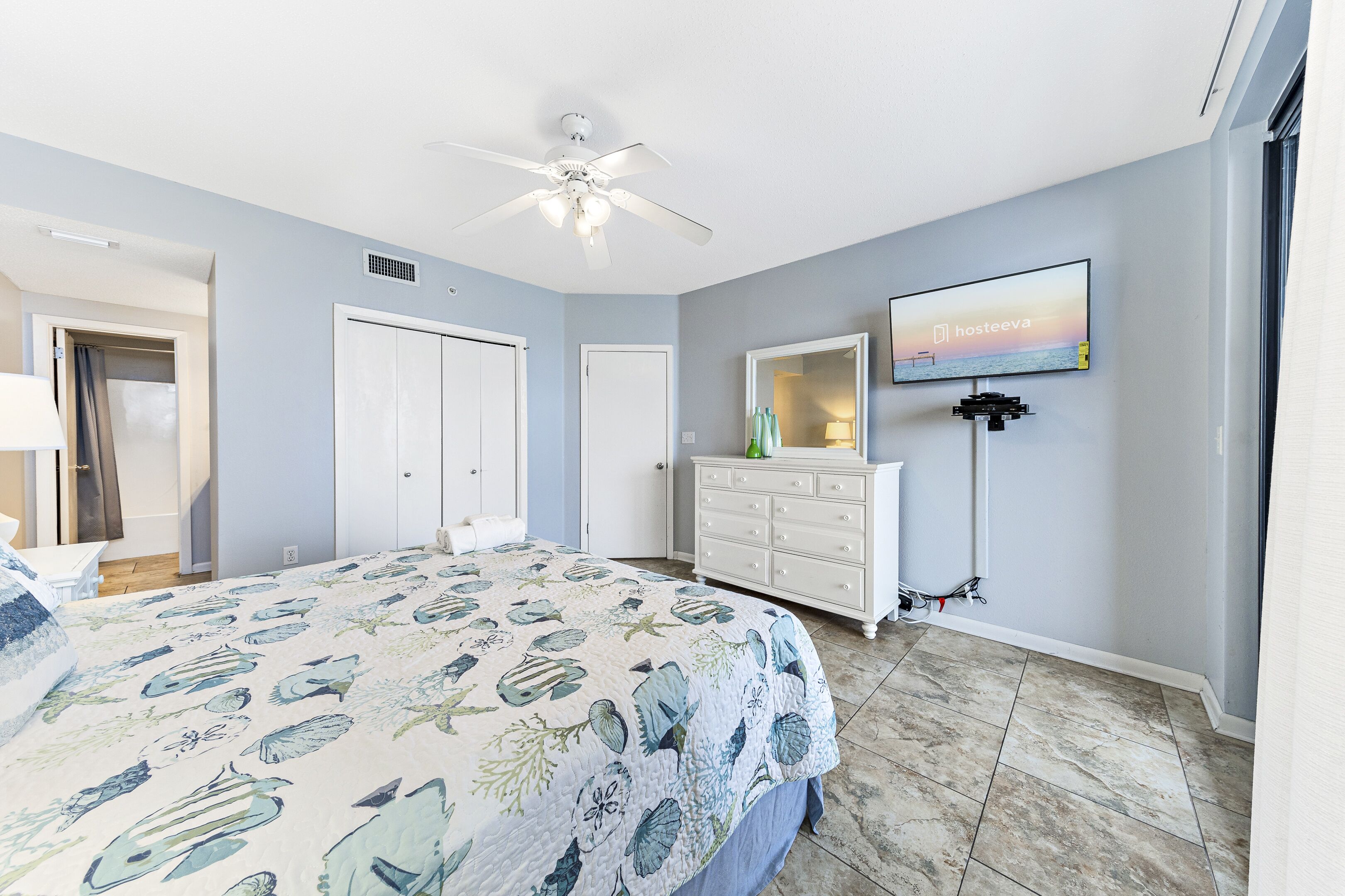 A bedroom with a bed, a ceiling fan, a dresser, a TV, and a door.