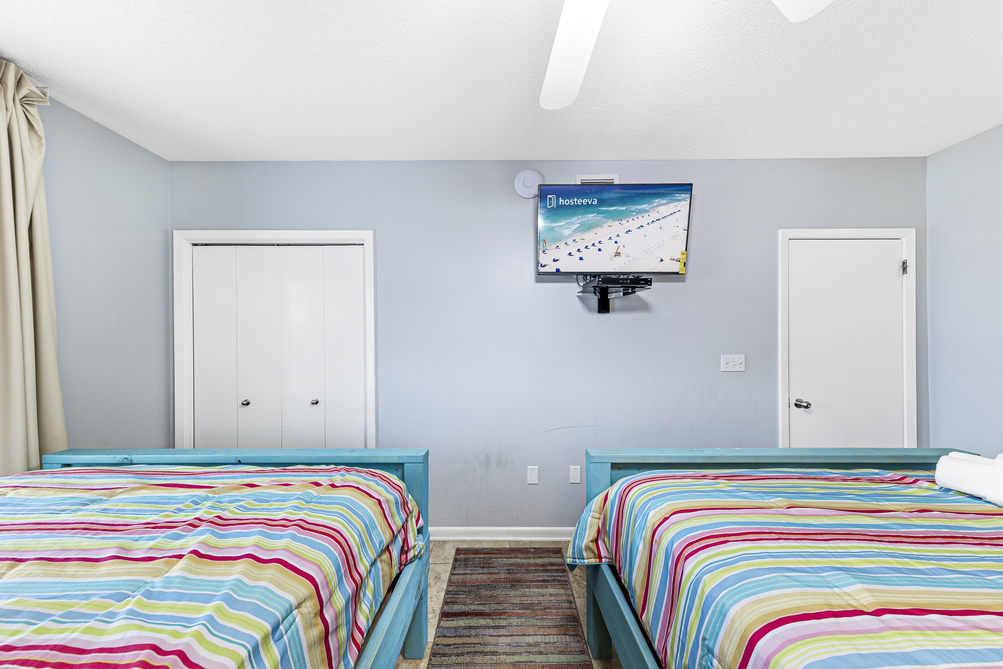A bedroom with two beds, a TV mounted on the wall, and a window with curtains.