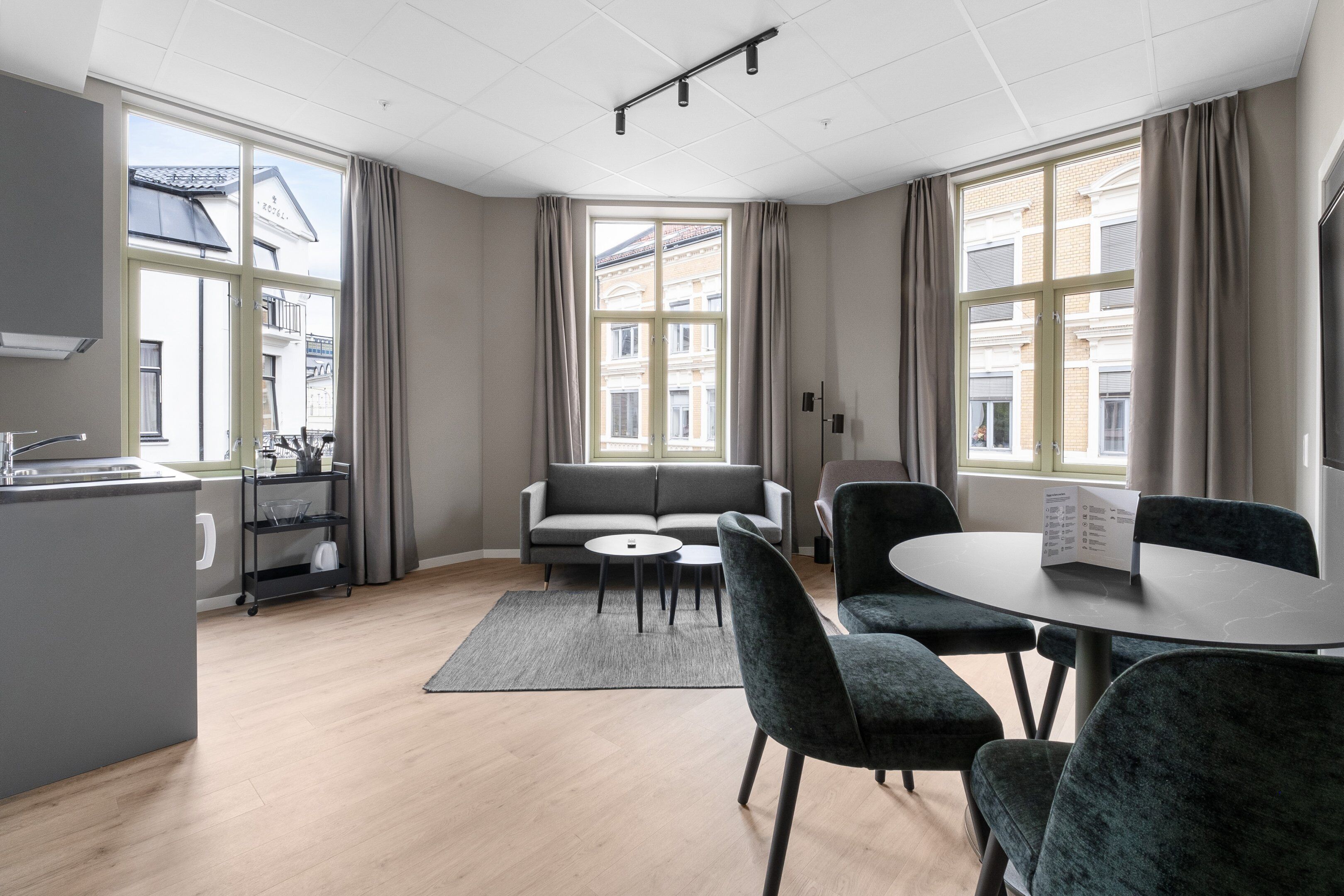 A modern hotel room with a kitchen, a gray sofa, a dining table with chairs, and large windows with curtains.