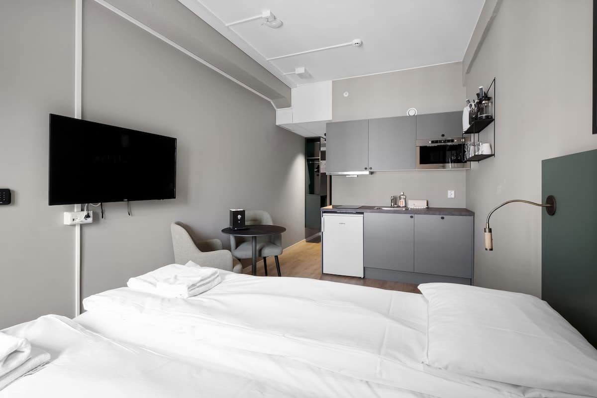 A modern hotel room with a bed, a desk, a chair, a small kitchenette, and a TV mounted on the wall.
