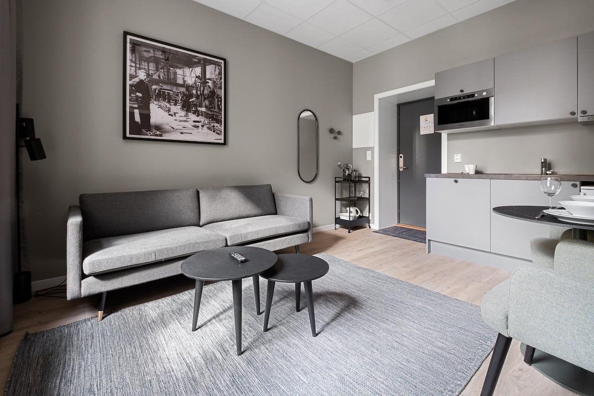 A modern living room with a grey sofa, two small round tables, a grey rug, a kitchen area with grey cabinets, and a black and white photograph on the wall.