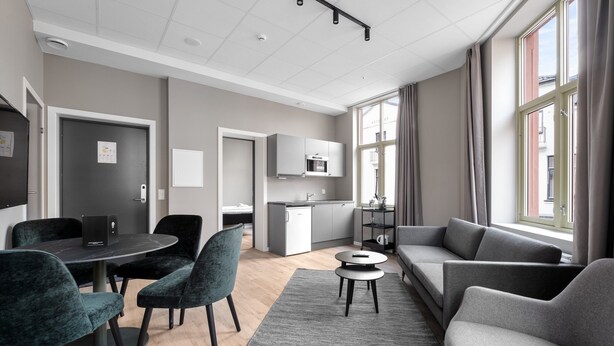 A modern living room with a grey sofa, a dining table with chairs, a kitchen area with cabinets, and a large window with curtains. - Forenom Serviced Apartments Drammen (Drammen)