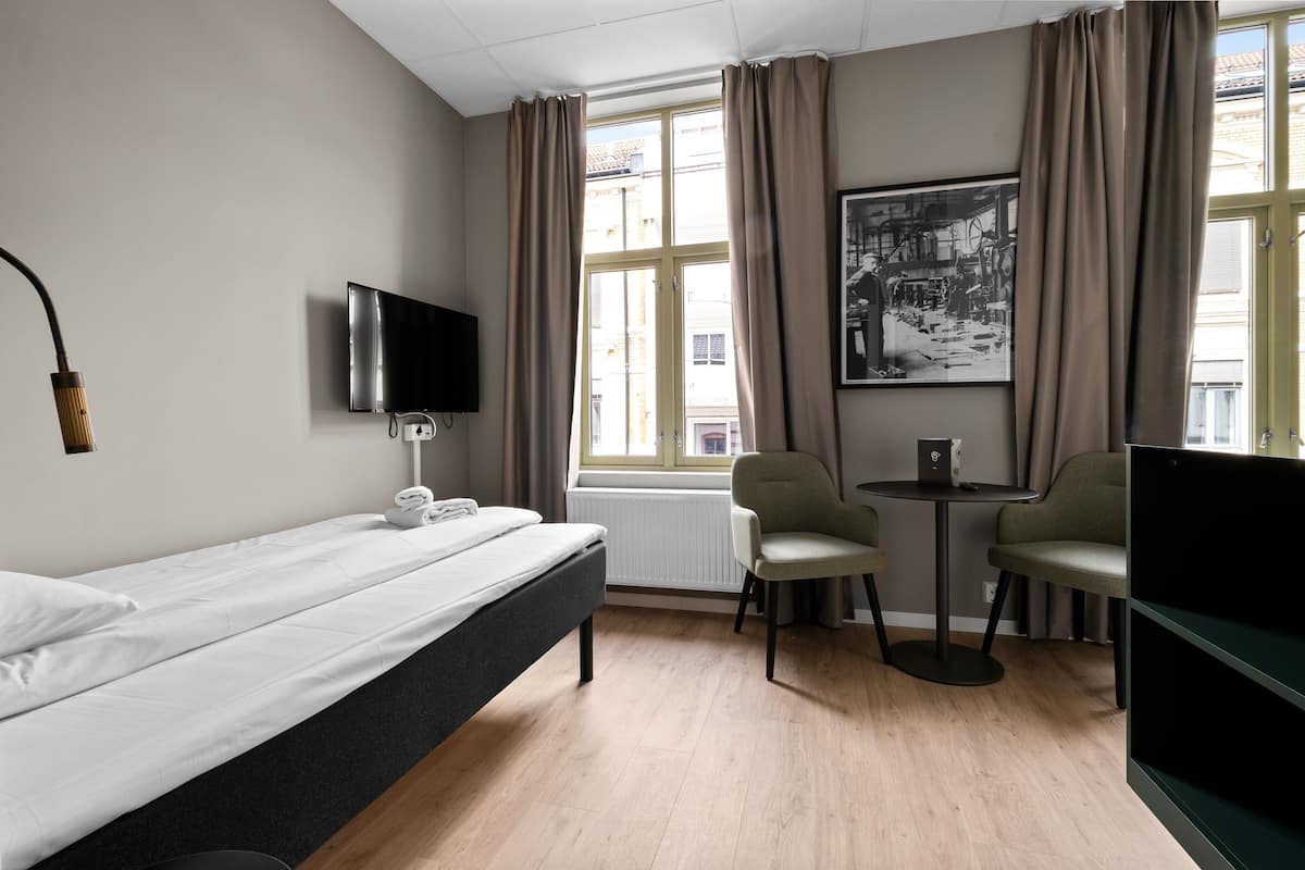 A hotel room with a bed, a desk with a laptop, two chairs, a TV, and a window with curtains.