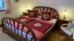 Ein gemütliches Schlafzimmer mit einem hölzernen Bett, rot-karierter Bettwäsche, herzförmigen Kissen, einem Nachttisch mit Lampe und einem Sessel mit Sitzkissen.