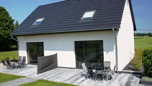 Ein modernes Haus mit geneigtem Dach, großen Schiebeglastüren und einer Terrasse mit Gartenmöbeln.