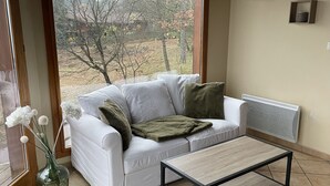 A white sofa with a green throw blanket and a small table in front. A vase with white flowers is on the floor beside the sofa. - L'Escapade Ardéchoise (Fons)