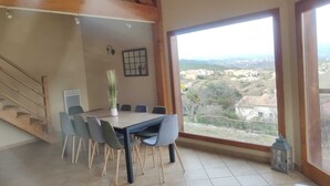 A dining area with a table and chairs, a view of a valley through large windows, and a staircase leading to an upper level. - L'Escapade Ardéchoise (Fons)