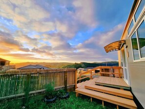 Um deck de madeira com um banco, um quintal cercado e vista para colinas distantes sob um céu parcialmente nublado.