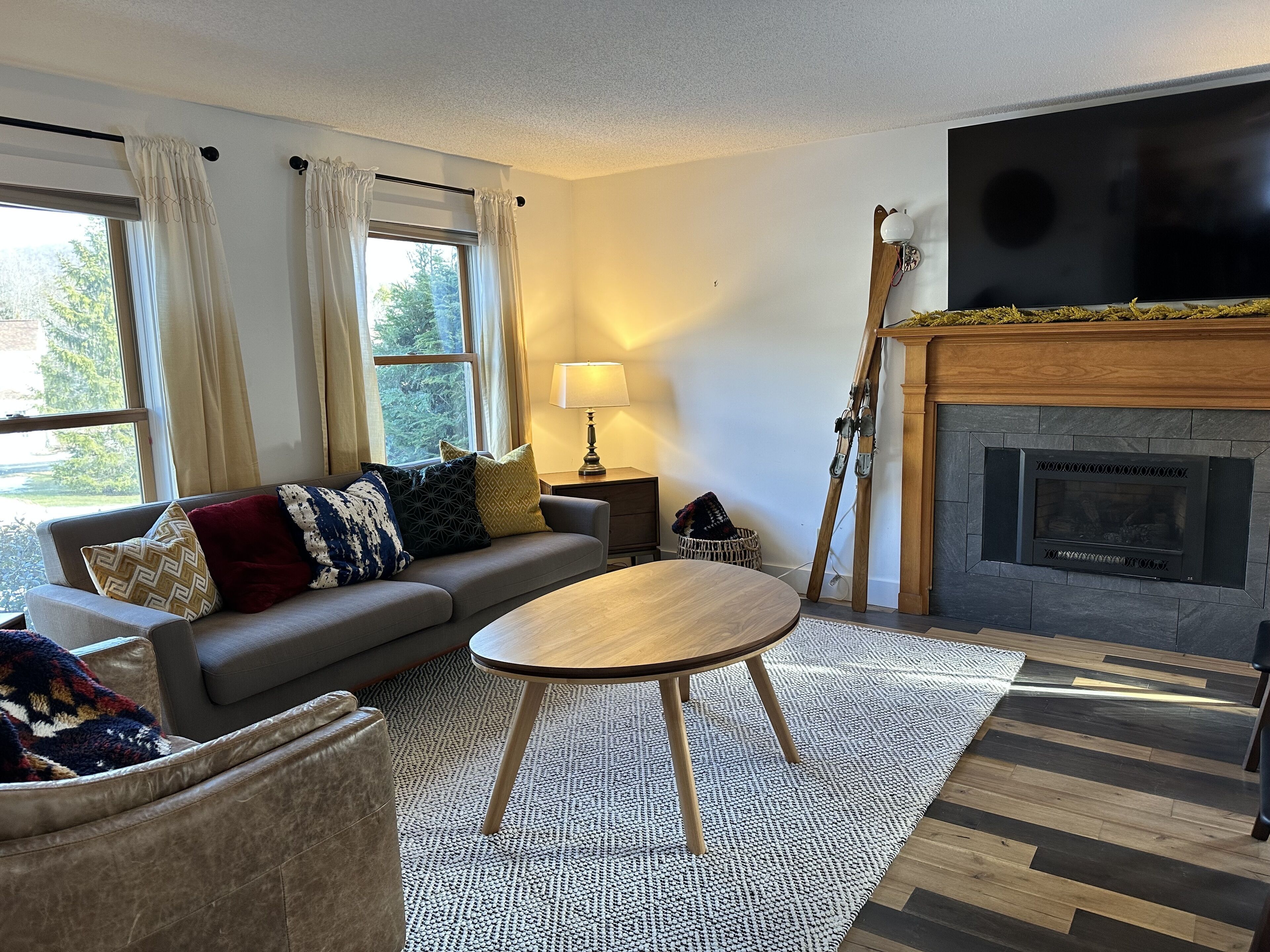 A living room with a sofa, a round wooden table, a fireplace, and a view of the outdoors through a window.