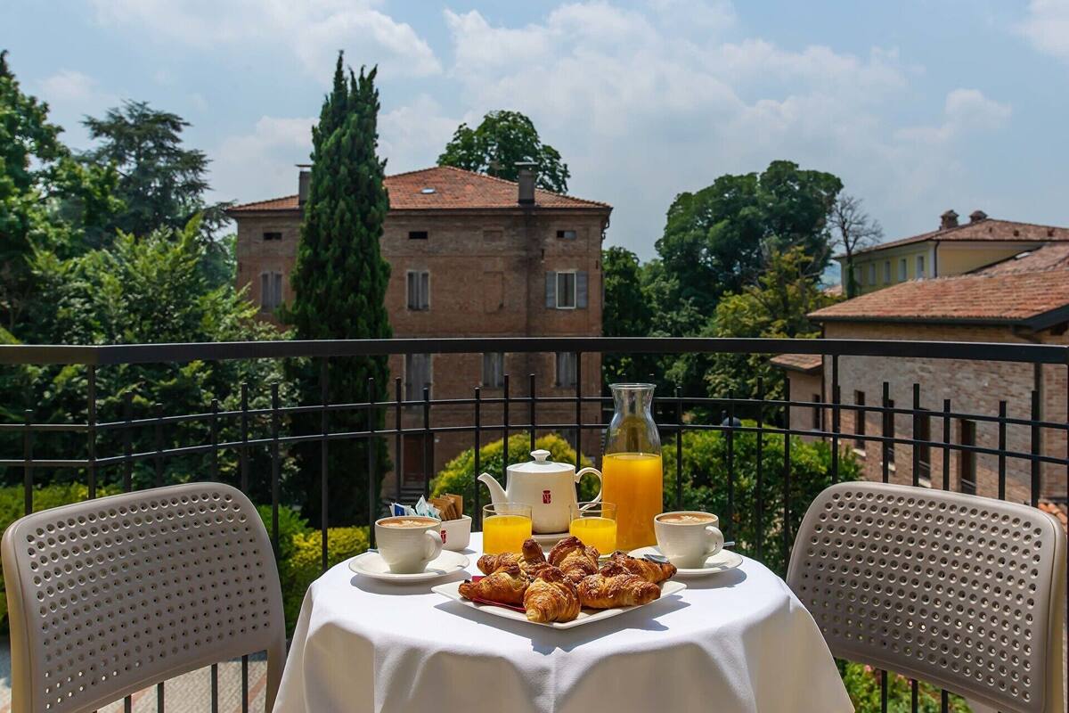 A table set with croissants, coffee, and orange juice on a balcony with a view of a brick building and trees.