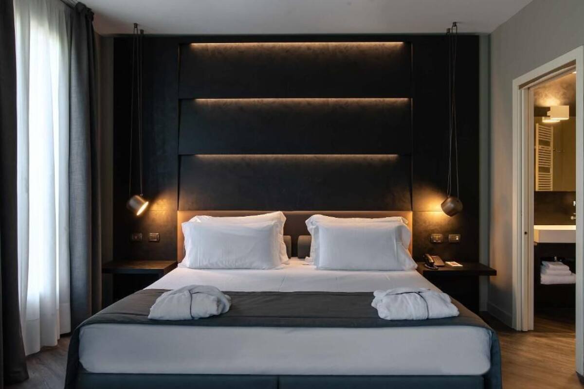 A modern hotel room with a large bed, bedside lamps, and a dark headboard.