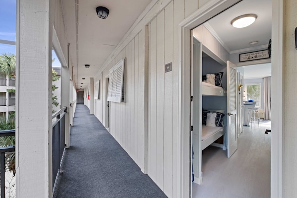 A corridor with a bunk bed room on the right, a balcony on the left, and a sign reading "Life Happens the Hard Way". - Just Beachy, Ocean View, steps to the beach, and Walking to Shopping and Dining🏖️ (Hilton Head Island)