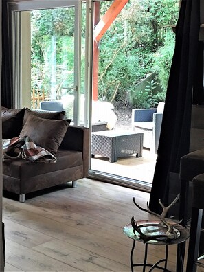 A modern living room with a sofa, a small table, and a glass door leading to an outdoor area with greenery.