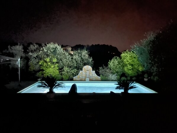 A nighttime view of a swimming pool with a stone fountain and lush greenery.