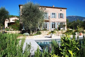 A two-story house with a swimming pool, surrounded by greenery and a mountain in the background.