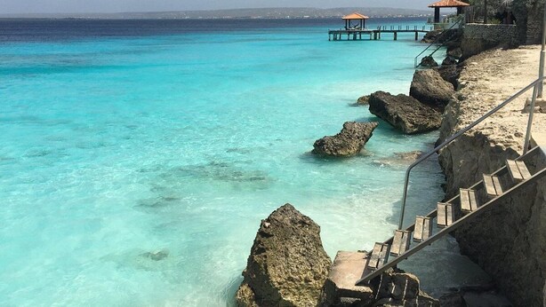 A coastal scene with clear turquoise water, rocky shoreline, and a wooden staircase leading down to the beach. - 2 Bed 1.5 Boutique home w pool, with Seaview deck 100 yrds from Bachelor's Beach (Belnem)