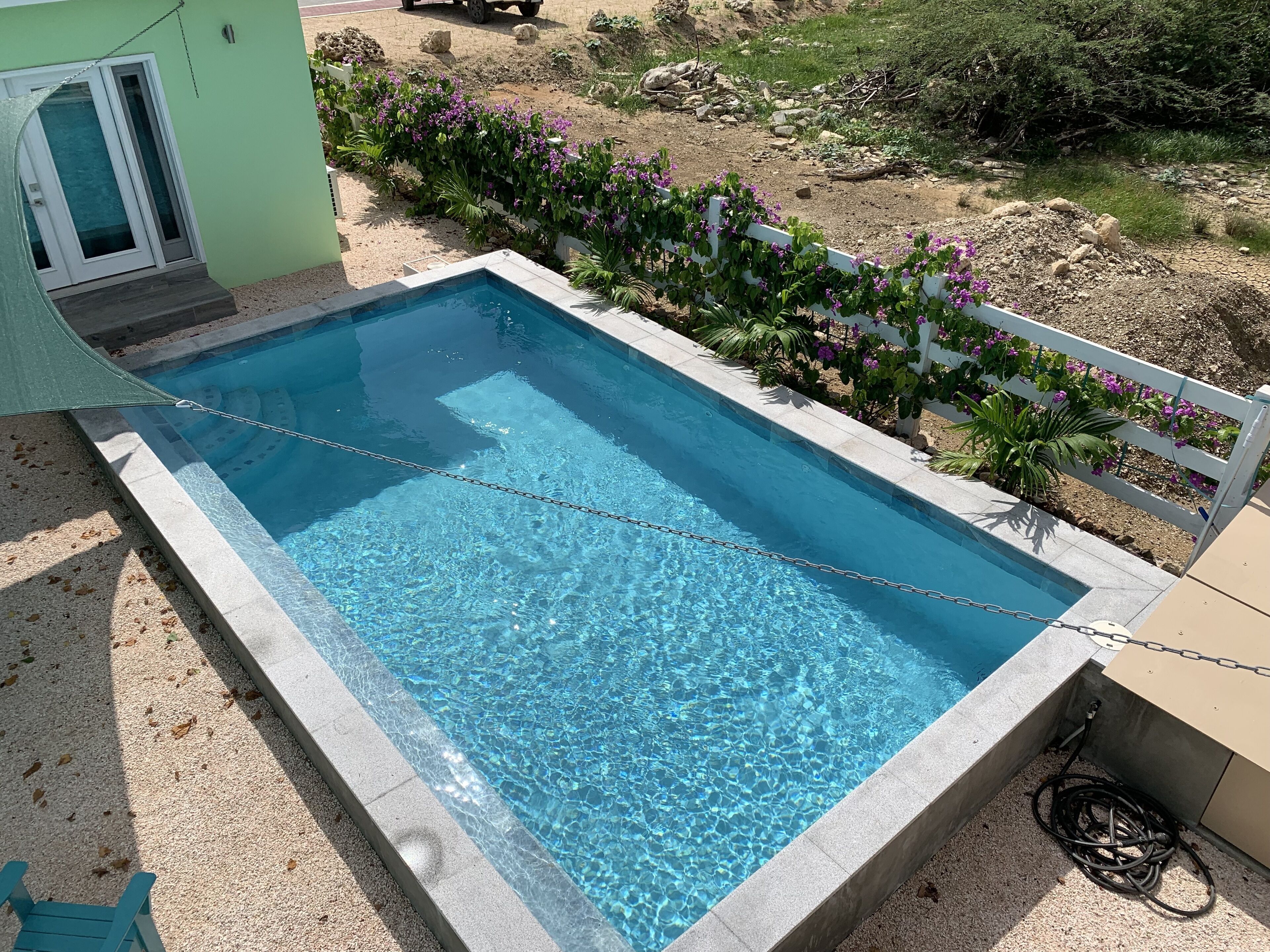 A rectangular swimming pool with clear blue water, surrounded by a white fence and purple flowers.