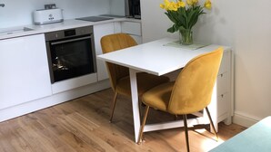 A compact kitchen with white cabinets, a toaster, and a coffee maker. There is a dining table with two yellow chairs and a vase of yellow flowers. A framed picture of a bird hangs on the wall. - A Newly Refurbished Apartment in Notting Hill, London (London)