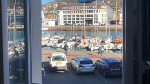 Blick aus einem Fenster auf einen Yachthafen mit Booten, ein Gebäude mit Beschilderung und eine hügelige Landschaft im Hintergrund.