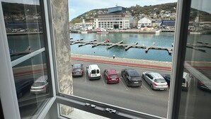 A view from a window overlooking a harbor with parked cars, a boat, and buildings in the background.